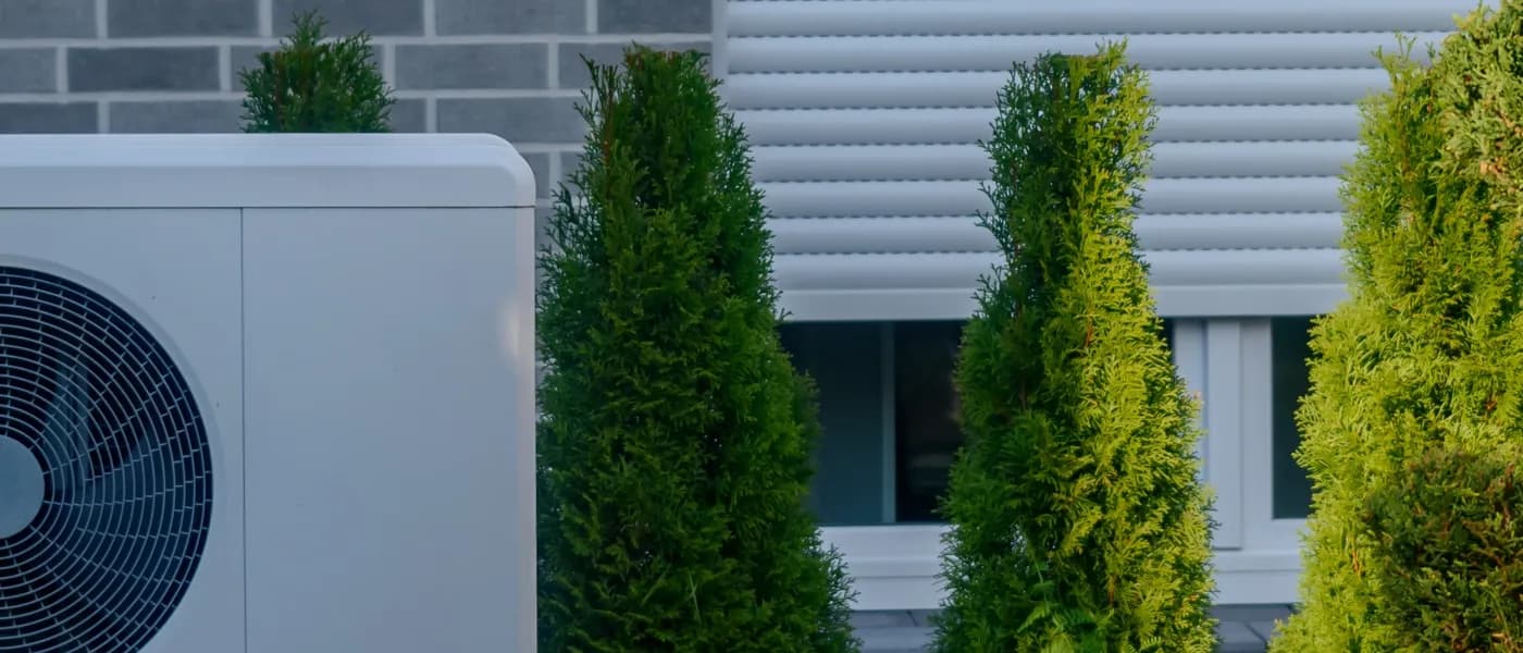 White outdoor heat pump unit installed next to evergreen bushes and a brick house wall.